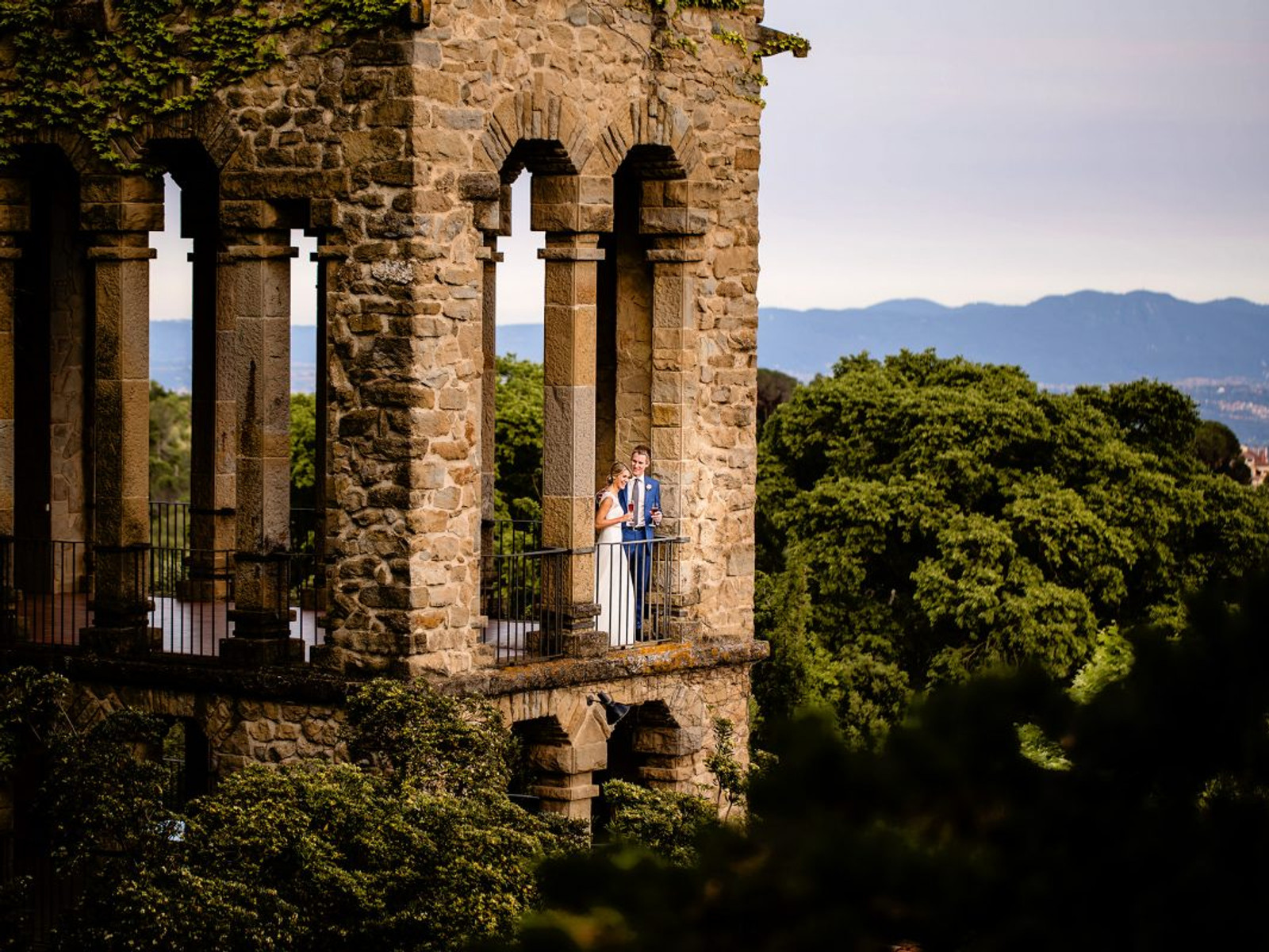A bride and groom sharing a toast on the iconic stone balcony of La Baronia, featuring Gothic arches and a panoramic view of the Catalan mountains near Barcelona.
